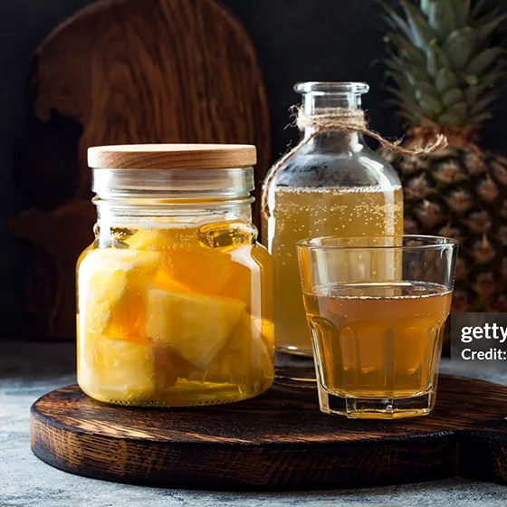 An image of a jar of Tepache next to a glass and bottle on a wooden board