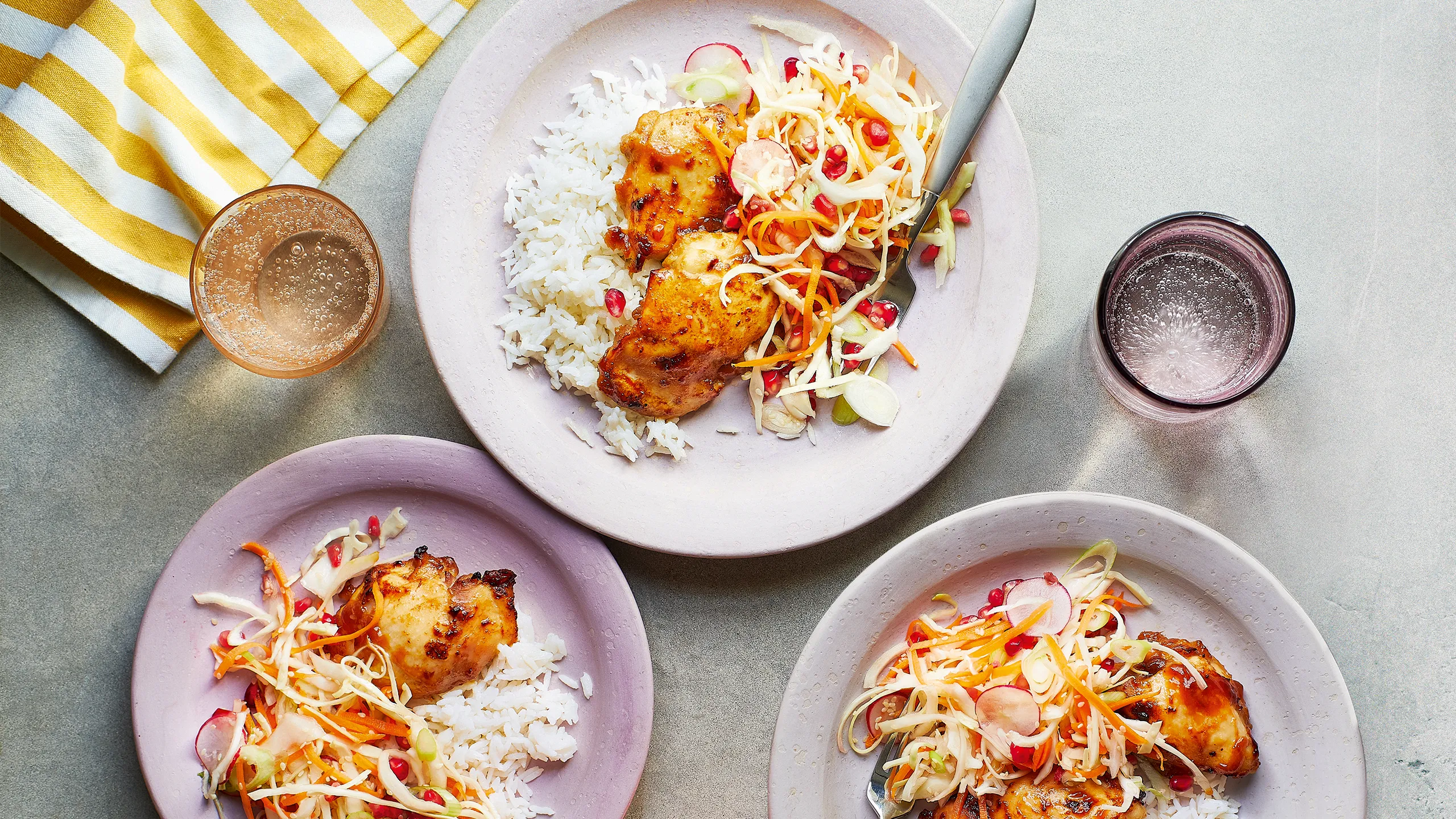 Three plates of grilled chicken on rice with vegetable slaw, accompanied by two glasses, on a light surface next to a yellow-striped cloth.