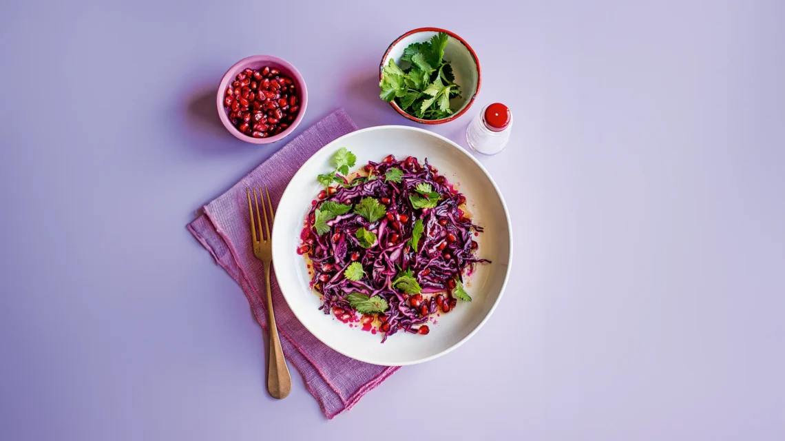Purple cabbage salad with pomegranate and cilantro on a lavender surface.