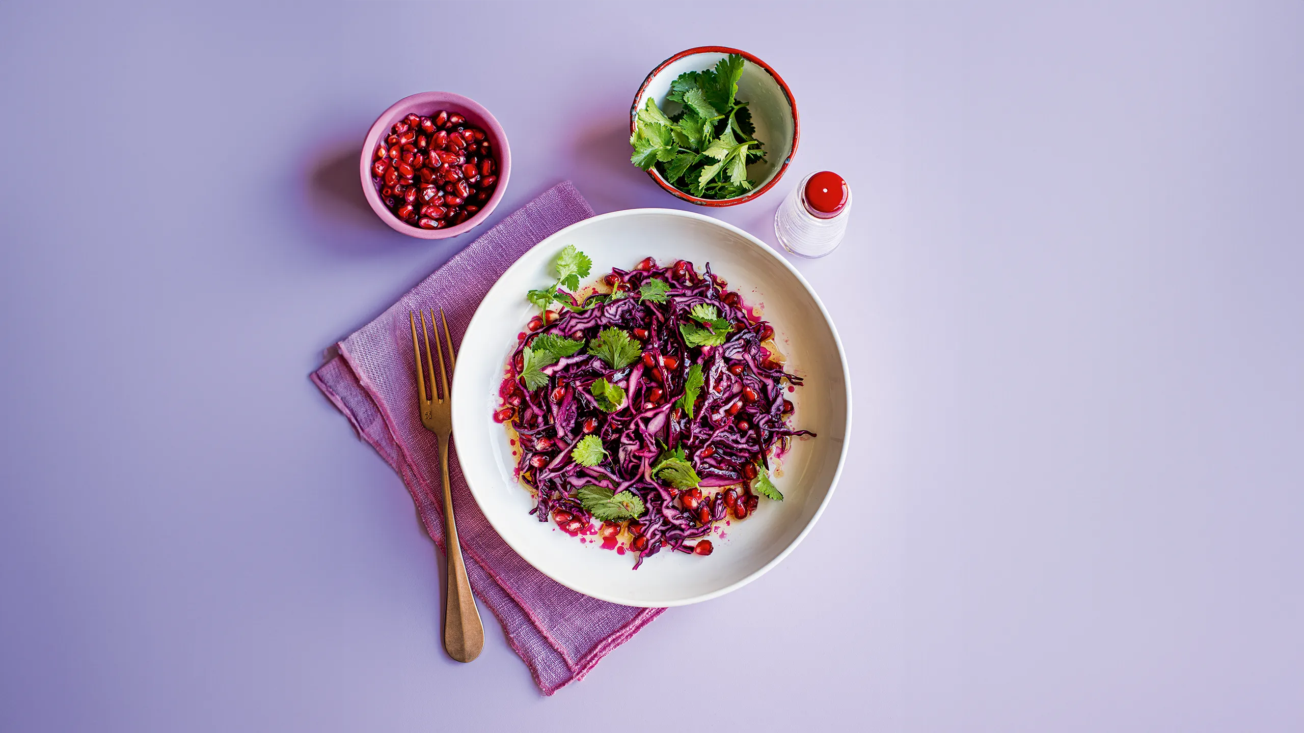 Purple cabbage salad with pomegranate and cilantro on a lavender surface.