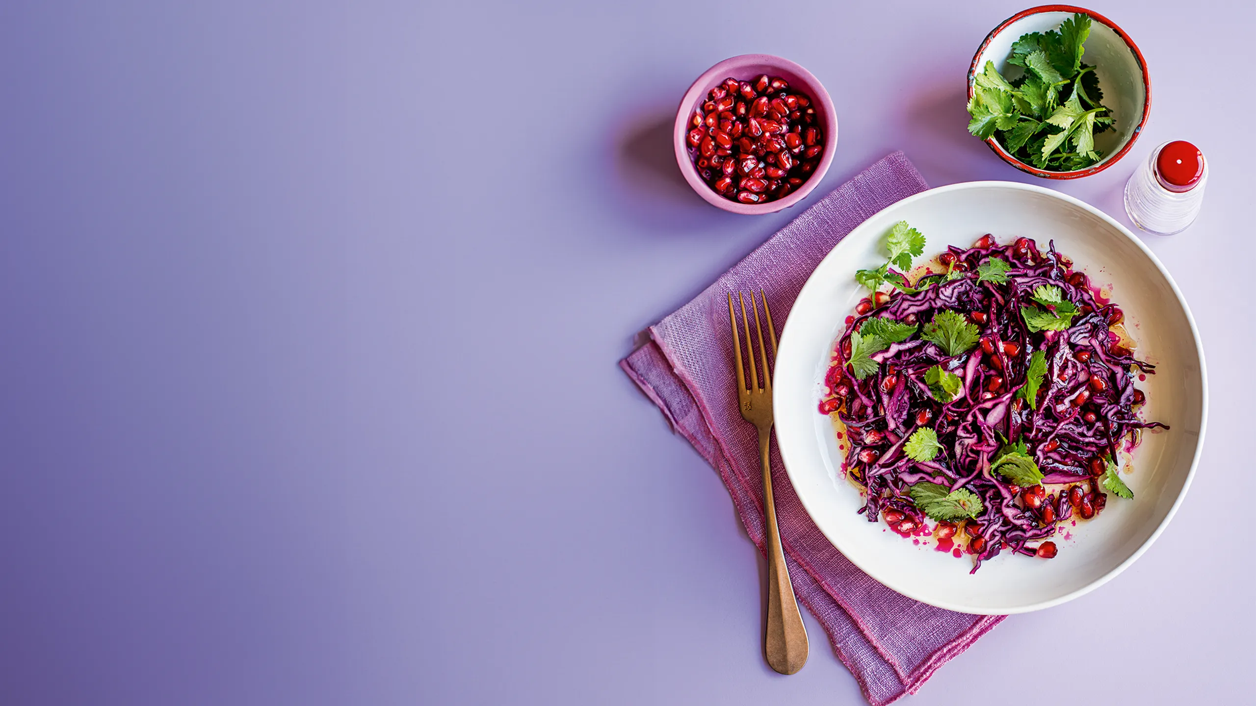 Purple cabbage salad with pomegranate and cilantro on a lavender surface.
