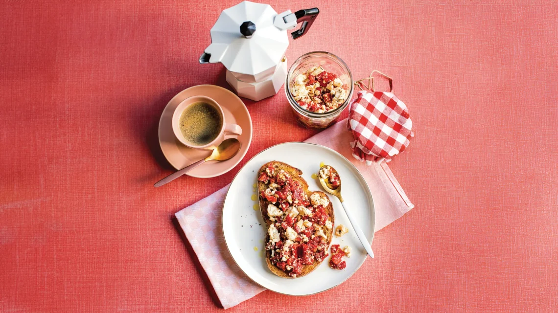 Toast with fruit and crumbles on a plate, jar with checked lid, coffee cup, and stovetop espresso maker on a red surface.