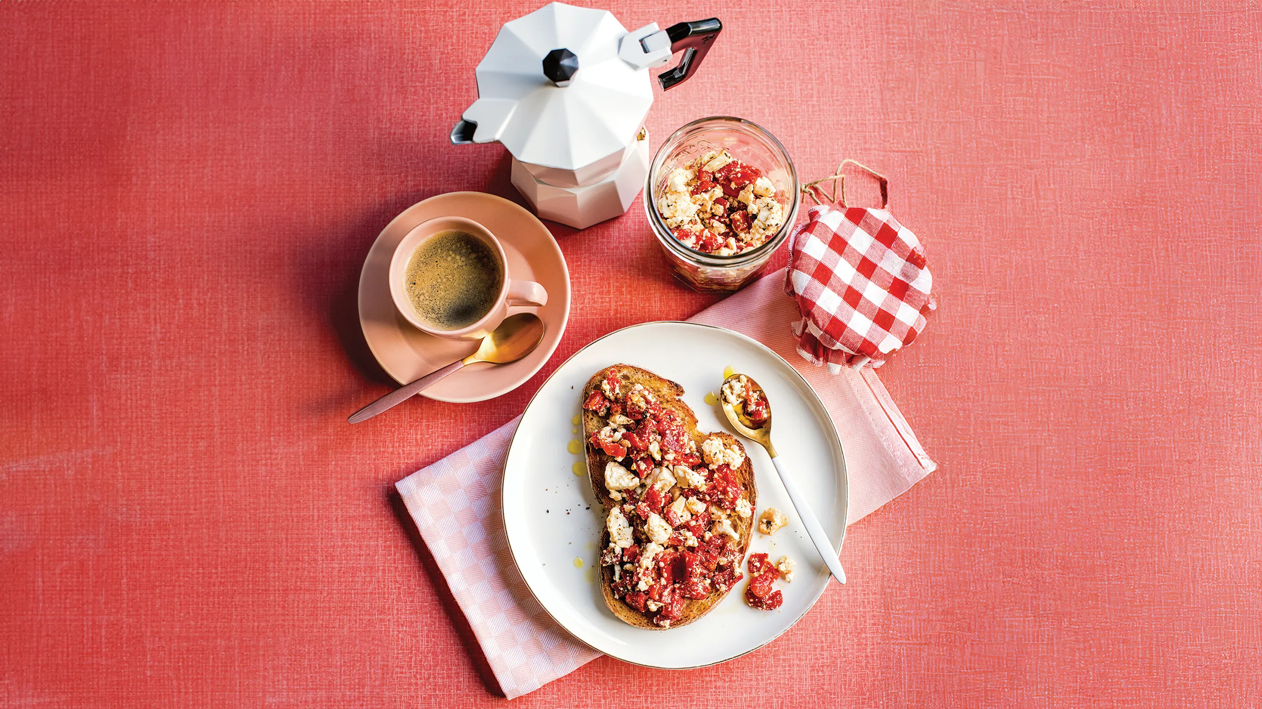 Toast with fruit and crumbles on a plate, jar with checked lid, coffee cup, and stovetop espresso maker on a red surface.