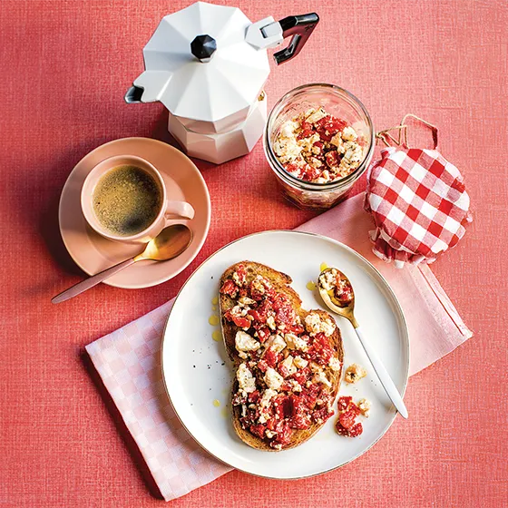 Toast with fruit and crumbles on a plate, jar with checked lid, coffee cup, and stovetop espresso maker on a red surface.