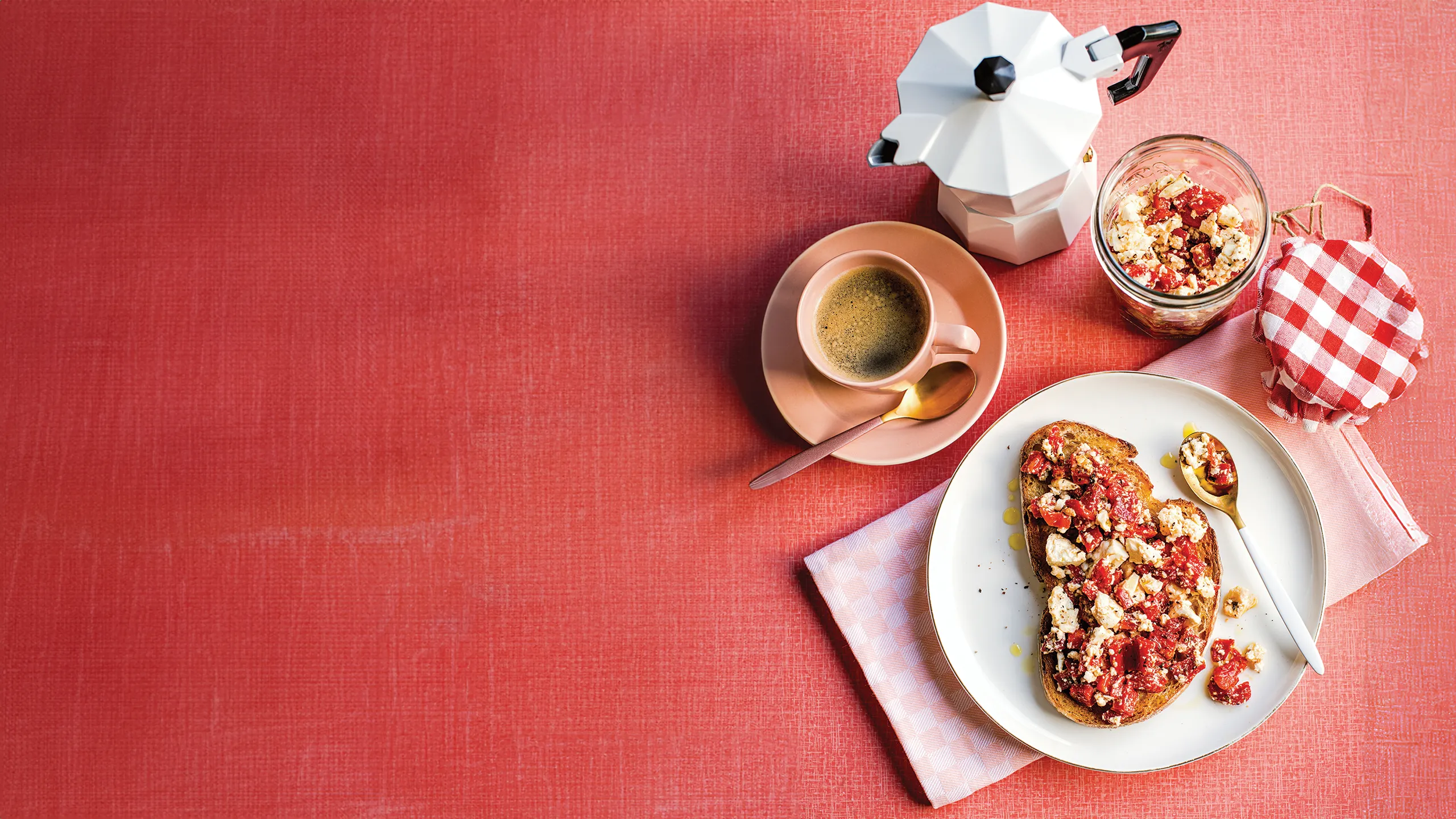 Toast with fruit and crumbles on a plate, jar with checked lid, coffee cup, and stovetop espresso maker on a red surface.