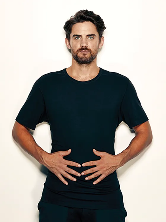Portrait of Stuart Sanderman on a light backdrop with his hand on his stomach area