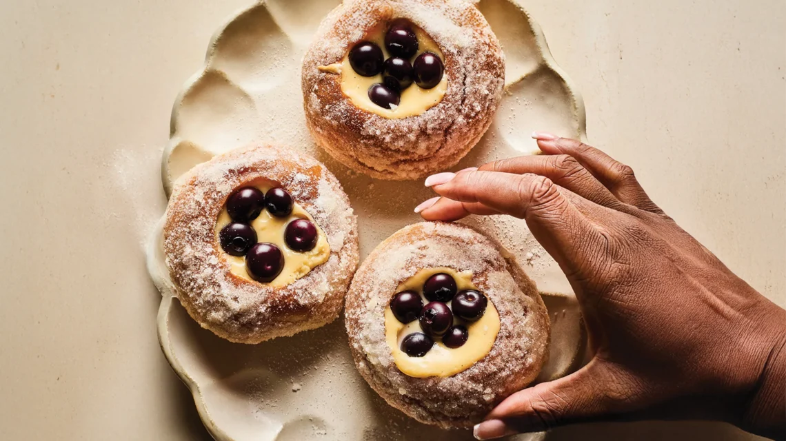 Three sugar-dusted doughnuts filled with cream cheese and topped with blueberries on a scalloped plate, as a hand reaches for one.