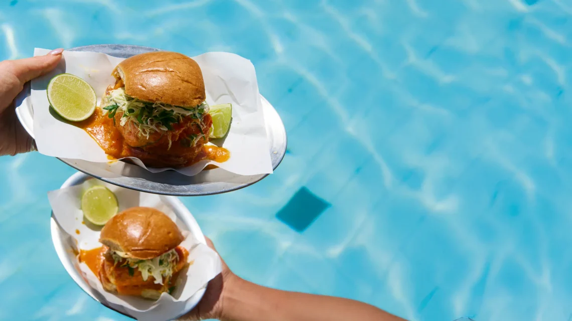 Two Moqueca burgers with lime wedges held on metal trays over the bright blue water of a swimming pool.