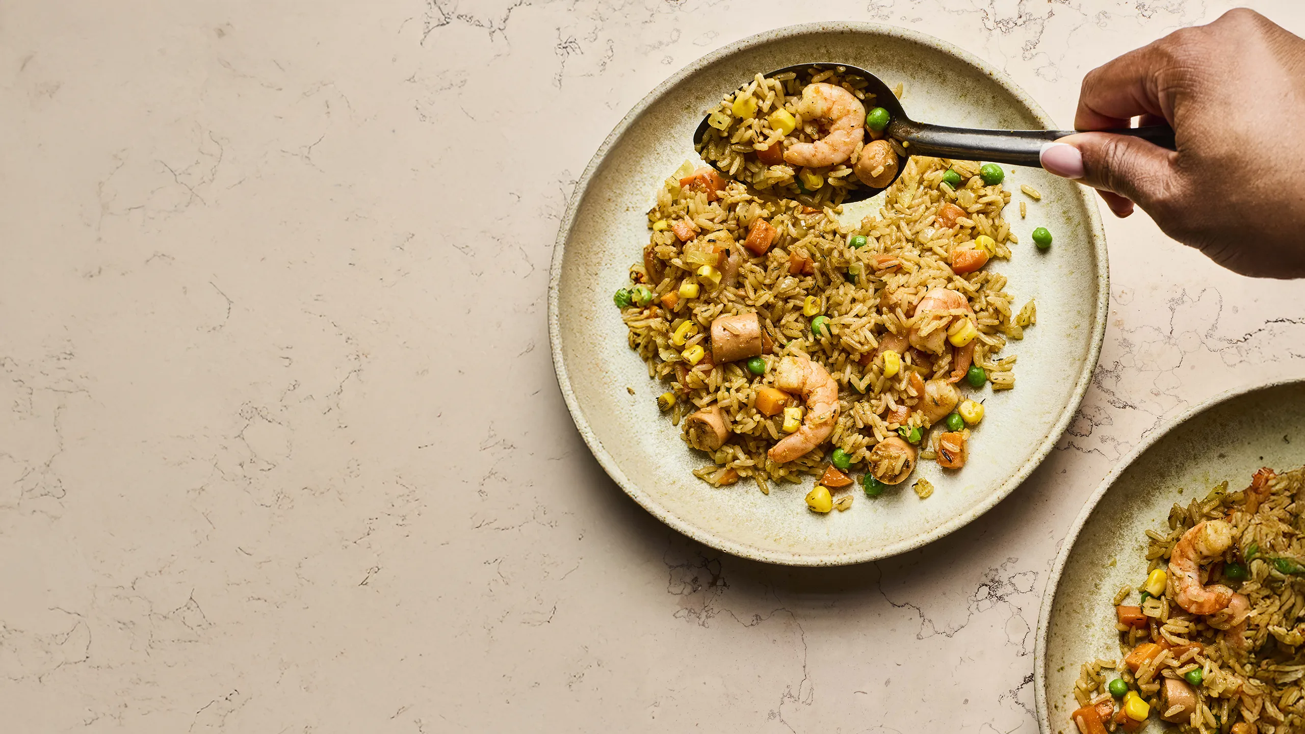 A person uses a spoon to serve West African fried rice mixed with prawns, sausage, and vegetables onto a stone plate.
