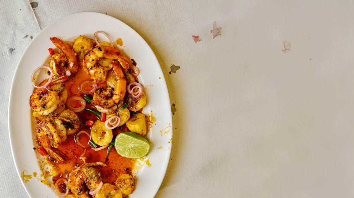 piced prawns and fried plantain served on a white platter with red onion rings, chillies, and a lime wedge.
