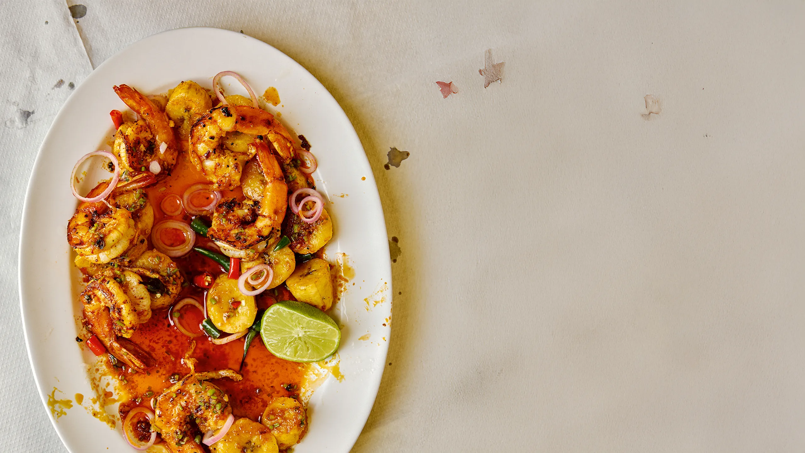 piced prawns and fried plantain served on a white platter with red onion rings, chillies, and a lime wedge.