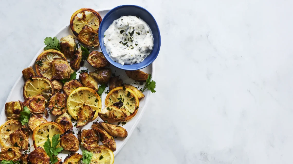 Roasted artichokes and charred lemon slices served with a small blue bowl of nori-flecked dip on a marble background.