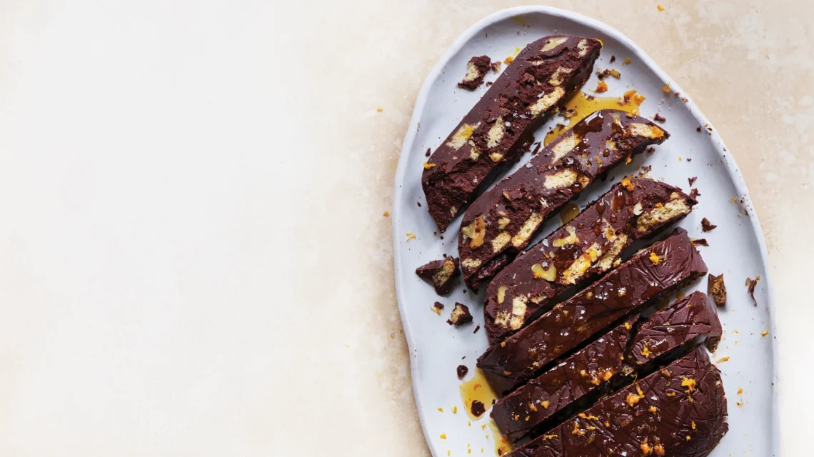 Slices of a chocolate tiffin or fridge cake with biscuit pieces, topped with orange zest and syrup on a grey platter.