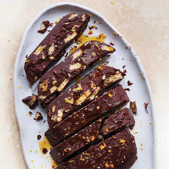 Slices of a chocolate tiffin or fridge cake with biscuit pieces, topped with orange zest and syrup on a grey platter.