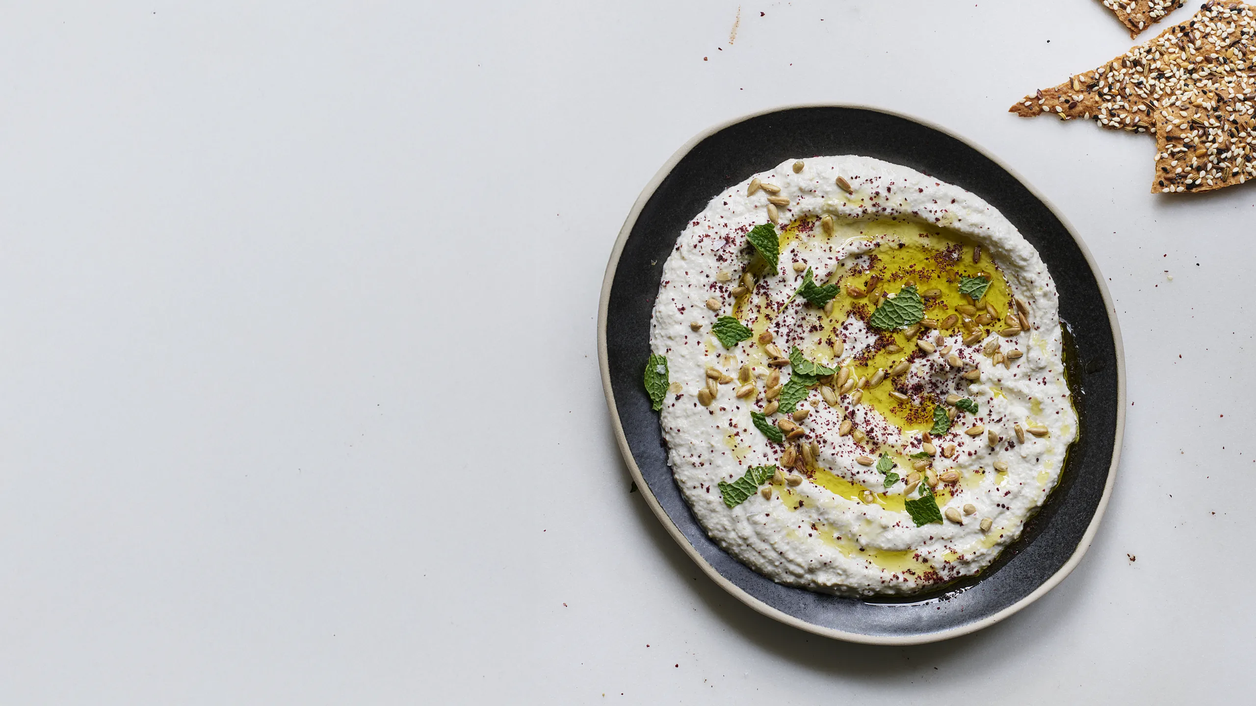 Creamy labneh topped with olive oil, sunflower seeds, and fresh mint, served on a dark plate with seeded crackers.