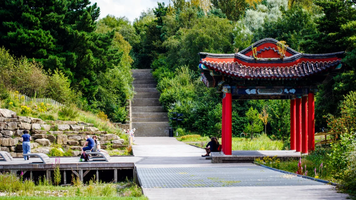 A traditional red Chinese pagoda stands beside a paved walkway in Liverpool Festival Gardens, with stone steps rising into the trees.