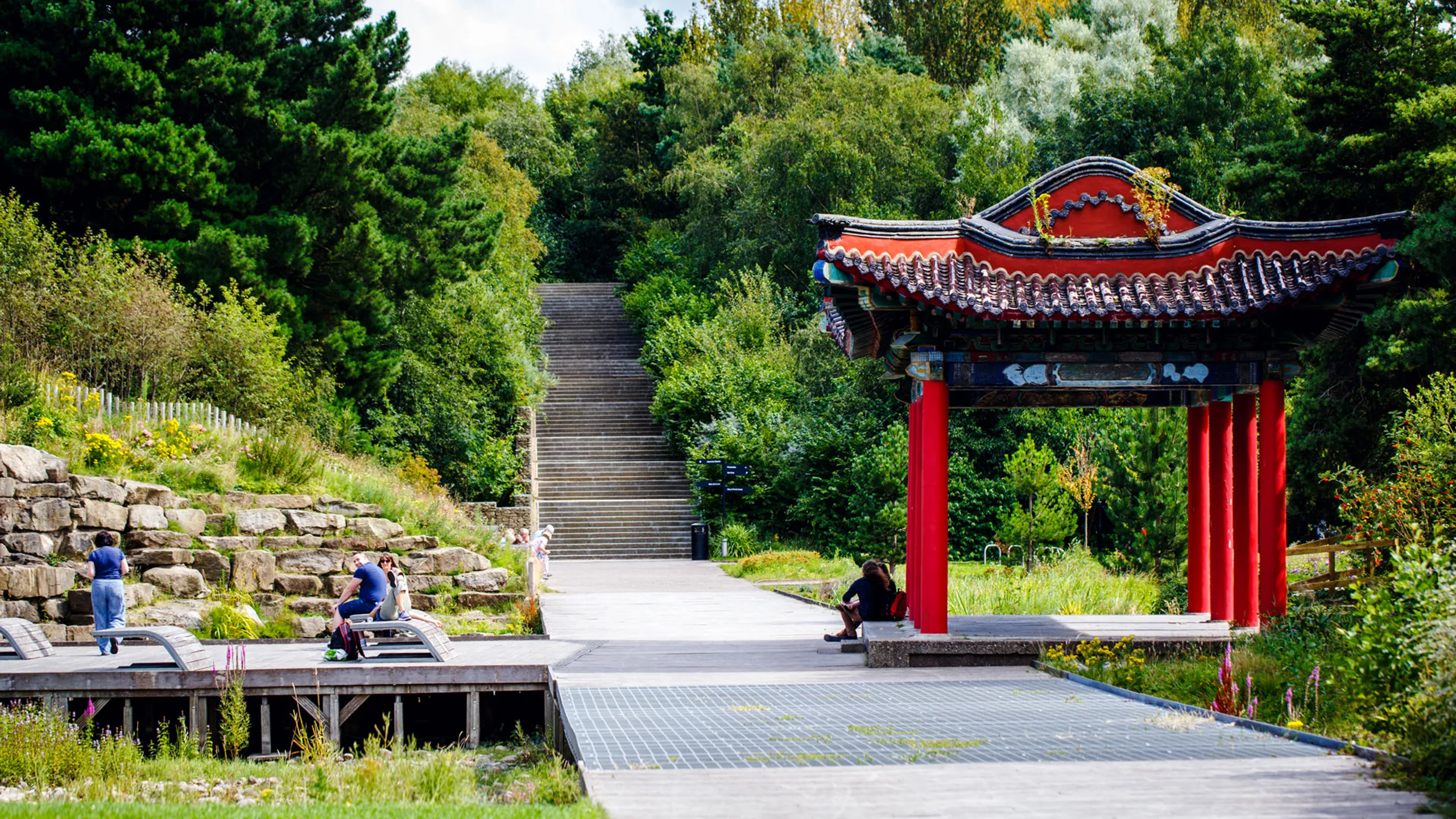 A traditional red Chinese pagoda stands beside a paved walkway in Liverpool Festival Gardens, with stone steps rising into the trees.