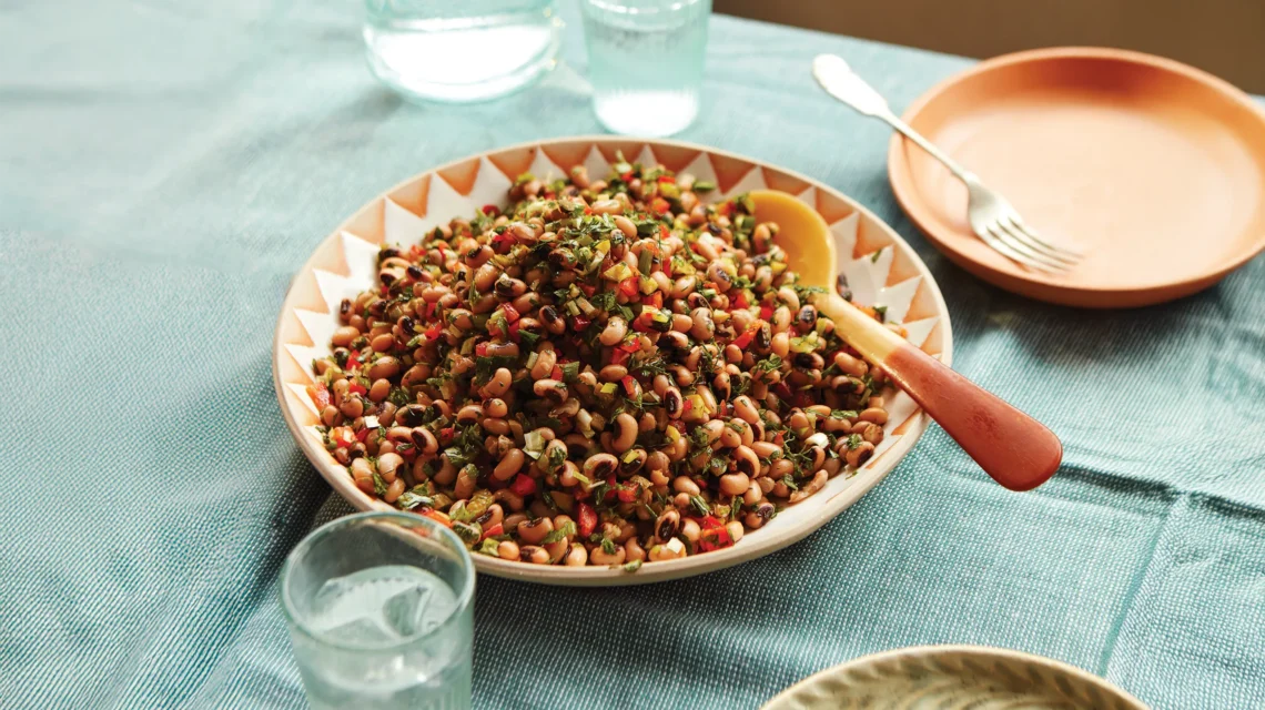 A patterned bowl filled with black-eyed bean salad mixed with herbs and peppers on a blue tablecloth.