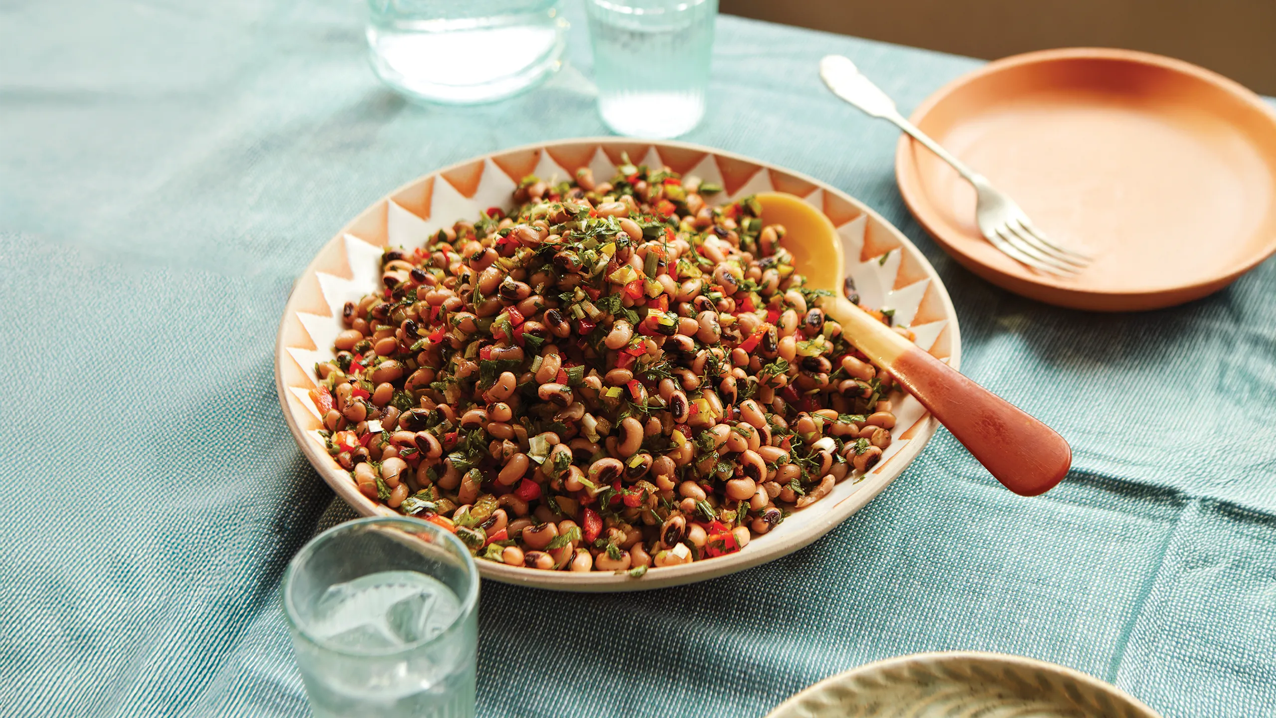 A patterned bowl filled with black-eyed bean salad mixed with herbs and peppers on a blue tablecloth.