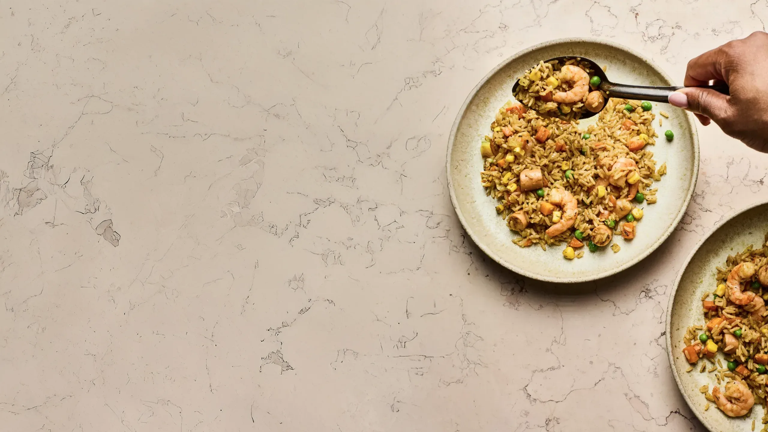 A person’s hand uses a black spoon to scoop West African fried rice with shrimp, peas, and carrots from a ceramic plate.