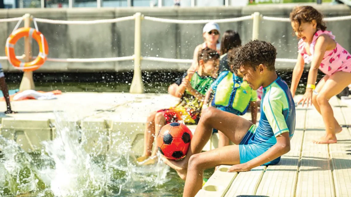 A group of children play on a floating dock; a boy balances a football on his foot while water splashes nearby.