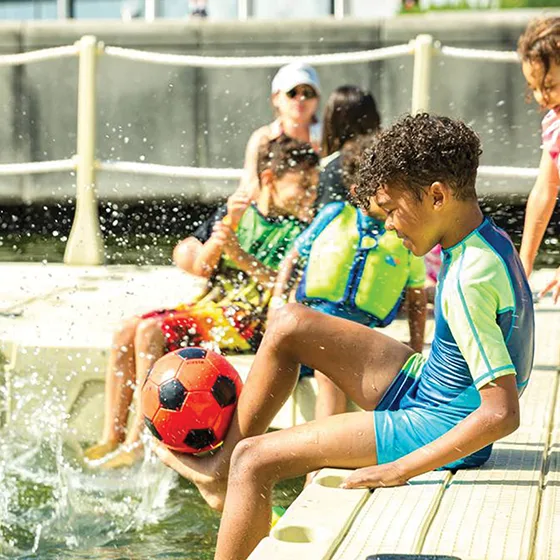 A group of children play on a floating dock; a boy balances a football on his foot while water splashes nearby.