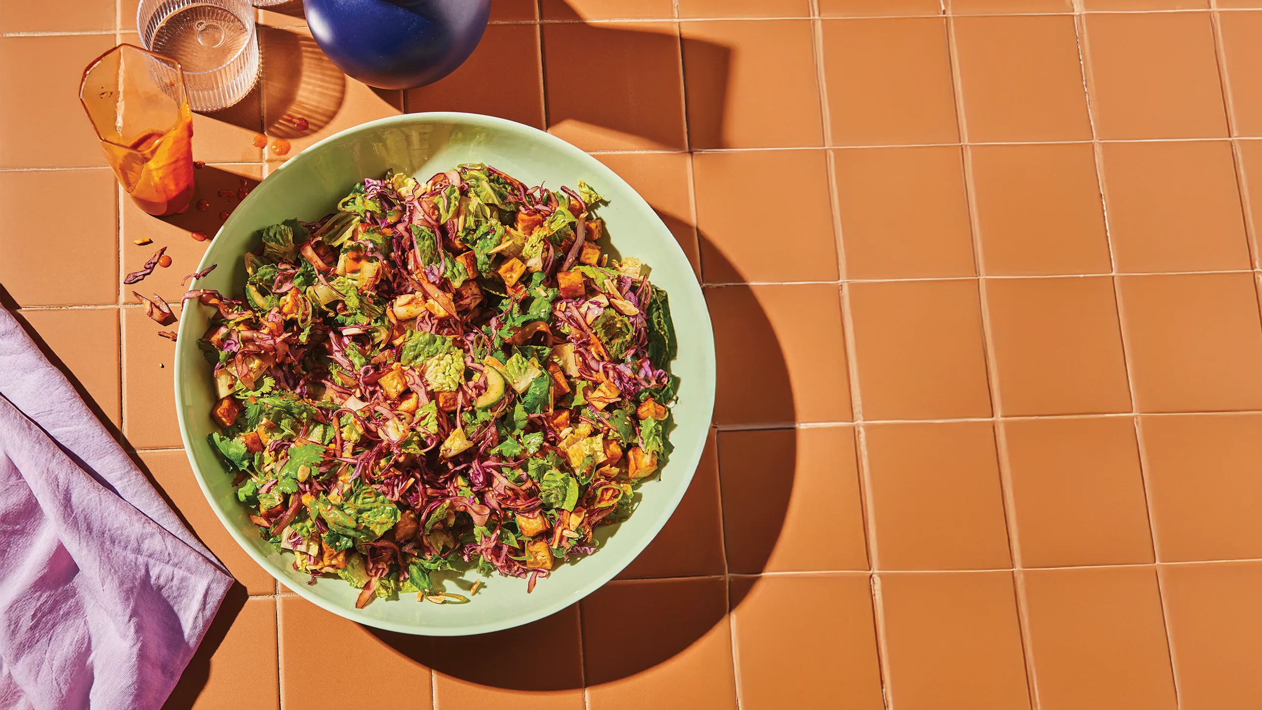 A large bowl of spicy peanut slaw with red cabbage, tofu croutons, and almonds on a terracotta tiled surface.