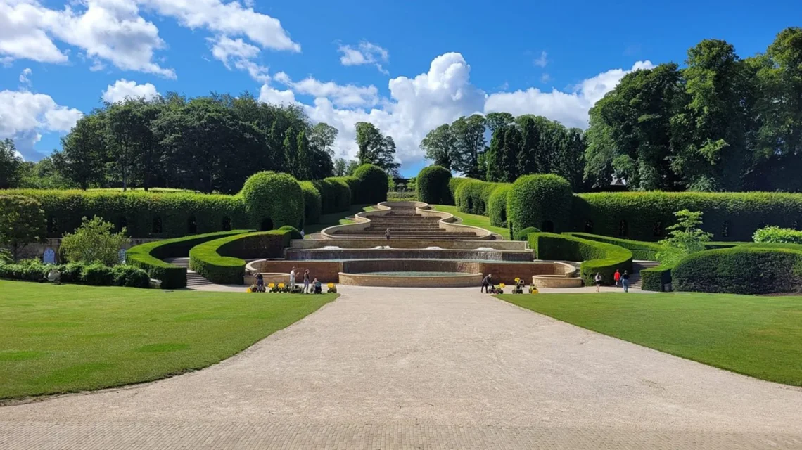 The Grand Cascade at The Alnwick Garden features tiered water fountains framed by sculpted hedges and a wide gravel path.