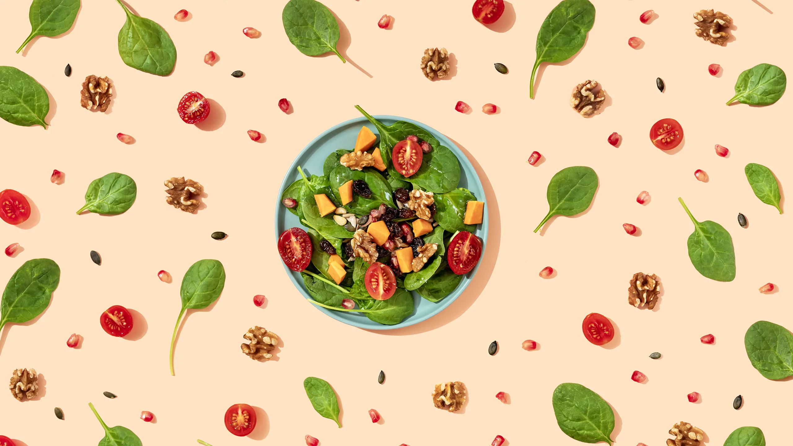 A flat lay of a vegan salad on a blue plate, surrounded by scattered spinach, walnuts, tomatoes, and pomegranate seeds.